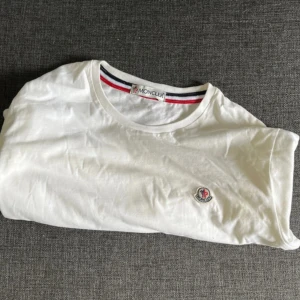 Vit Moncler t-shirt med logga - Vit t-shirt från Moncler med klassisk rund hals och diskret broderad logga på bröstet. Halsringningen har snygga ränder i rött, vitt och blått som ger en sportig touch. Tillverkad i mjuk bomull för maximal komfort. Priset går att diskutera 