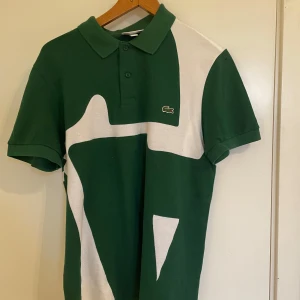 Grön och vit pikétröja från Lacoste - Snygg pikétröja från Lacoste i grönt och vitt med stor grafisk design och klassisk krokodillogga på bröstet. Tröjan har krage, knappar och korta ärmar. Regular fit och tillverkad i bomull, perfekt för en clean och sportig look. Om du är bajare passar den ännu bättre