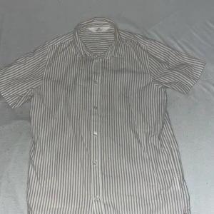 Randig kortärmad skjorta - Snygg kortärmad med beige och vita vertikala ränder. Skjortan har klassisk krage och knäppning framtill. Perfekt för en avslappnad och stilren look under varmare dagar.