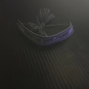 Dior Armband - Helt ny.Snyggt vävt armband från Christian Dior med texten 'J'Adior'. Armbandet är lila med mörkare broderad text och har flätade band med tofsar i ändarna. Perfekt accessoar för att lyfta din outfit med en lyxig touch.