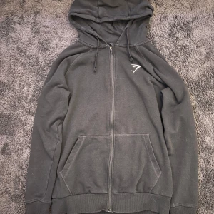 Svart hoodie från Gymshark med dragkedja - Svart hoodie från Gymshark med hel dragkedja, huva med snörning och två stora fickor framtill. Diskret Gymshark-logga på bröstet. Perfekt för chill eller träning, mjukt bomullsmaterial och klassisk passform.