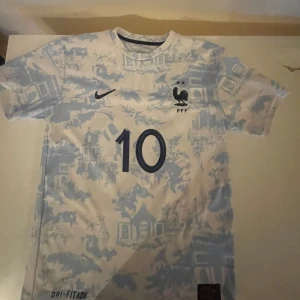 Frankrike Mbappé #10 fotbollströja Nike - Snygg fransk landslagströja från Nike med Mbappé och nummer 10. Tröjan är vit med ljusblått mönster, har korta ärmar och klassisk rund hals. Tryckta detaljer och Dri-Fit material som andas perfekt för fotboll eller träning.