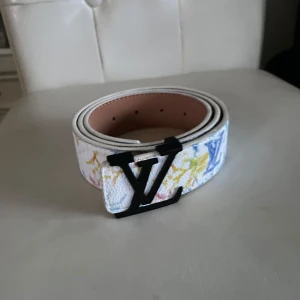 Vitt bälte med färgglada mönster - Säljer ett vitt bälte i skinn med färgglada  och mönster i blått, gult och rosa. Bältet har ett svart spänne framtill och är tillverkat i Spanien. Perfekt för dig som vill sticka ut med en lyxig accessoar.