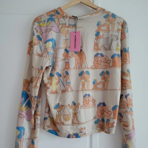 Beige långärmad topp Carne Bollente XL - Unik långärmad topp från Carne Bollente i beige med färgglatt, tecknat mönster inspirerat av antika motiv. Tillverkad i mjuk lyocellblandning med stretch. Rund halsringning och figurnära passform. Perfekt för dig som gillar statement-plagg och konstnärlig stil.
