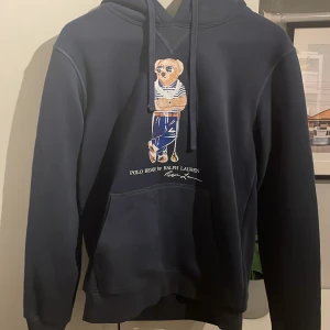 Mörkblå Polo Bear hoodie från Ralph Lauren - Säljer en mörkblå hoodie från Ralph Lauren med Polo Bear-tryck på bröstet. Tröjan är bara testad och lappar/etikett finns kvar i den. Klassisk känguruficka, huva med snörning och långa ärmar. Perfekt för dig som gillar streetstyle och vill ha något ikoniskt och bekvämt. Tröjan är Medium men mer åt small. Skriv vid minsta fundering!