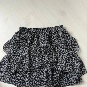 Svart blommig volangkjol - Supersöt svart kortkjol med vita små blommor och flera volanger. Kjolen har resår i midjan och är fodrad för extra komfort. Perfekt för dig som gillar en feminin och trendig stil.