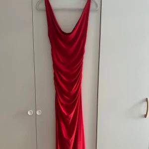 Röd långklänning med drapering - Snygg röd långklänning med draperad effekt och djup urringning både fram och bak. Klänningen har breda axelband och är figurnära med ett elegant fall. Perfekt för dig som vill sticka ut med en klassisk men ändå modern look