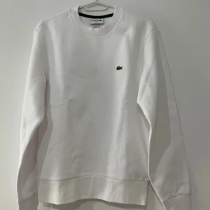 Vit Lacoste tröja - Klassisk vit sweatshirt från Lacoste med rund hals och broderad grön krokodil på bröstet. Tröjan har lång ärm och normal passform, tillverkad i mjuk bomull för en clean och stilren look. Perfekt för dig som gillar minimalistisk stil.