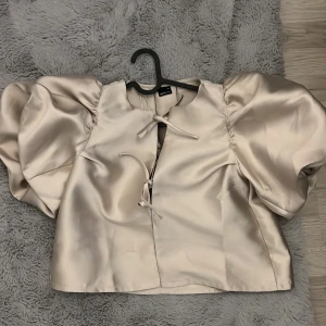 Champagnefärgad blus från Gina Tricot - Supersnygg champagnefärgad blus från Gina Tricot med puffiga ärmar och croppad passform. Blusen har knytning framtill och är gjord i ett glansigt satinliknande material som ger en lyxig känsla. Perfekt för dig som vill sticka ut med coola detaljer och volym.