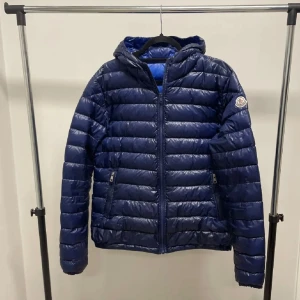 Moncler Dominic Jacka - Snygg blå dunjacka från Moncler med klassisk logga på ärmen. Jackan har horisontella sömmar, dragkedja framtill och två fickor med dragkedja. Utrustad med huva och lätt glansig finish som ger en modern vibe. Perfekt för kyliga dagar.