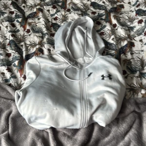 Vit hoodie från Under Armour - Säljer en vit hoodie från Under Armour med dragkedja och huva. Tröjan har snörning i huvan, två fickor framtill och en svart logga på bröstet. Perfekt för chill dagar eller när du vill ha en sportig look.