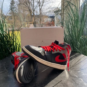 Nike Air Jordan 1 med röda detaljer - Hej!                                                                            Säljer nu mina Nike Air Jordans för dem har bara luggit i mitt garderob ett tag. Dem är svarta och röda med grå detaljer. Skorna har en klassisk hög modell med röd snörning och det ikoniska Air Jordan-märket på sidan. Perfekta för dig som vill ha en stilren och sportig look. Orginalpris är runt 1500kr.