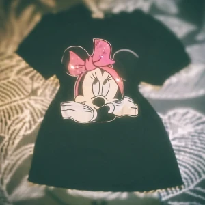 Svart t-shirt med Minnie Mouse-motiv - Säljer en svart t-shirt med ett stort tryck av Minnie Mouse på framsidan. Motivet har en söt rosa rosett med paljetter som ger en glittrig effekt. Perfekt för alla Disney-fans! 🖤