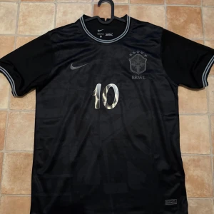 Svart Brasilien fotbollströja Nike #10 - Snygg svart Brasilien fotbollströja från Nike med silvriga detaljer och nummer 10 på både fram- och baksidan. Tröjan har korta ärmar, rund hals med grå ränder och är tillverkad i ett lätt och ventilerande material. Perfekt för dig som älskar fotboll och vill sticka ut.