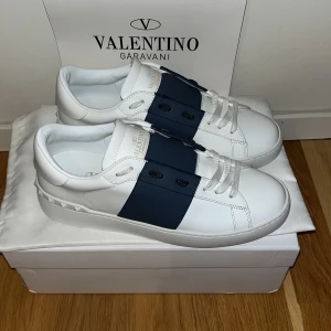 Valentino Garavani Open sneakers HELT NYAvit/marin - Snygga Valentino Garavani Open sneakers i vitt skinn med en bred marinblå läderdetalj över mitten. Klassisk rund tå, vita snören och platt sula. Perfekta för dig som vill ha en clean men ändå lyxig look. 