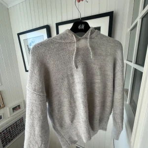 Ljusgrå stickad hoodie från Gina Tricot  - Supermjuk och mysig ljusgrå hoodie från Gina Tricot med huva och snörning. Tröjan är stickad med ribbade muddar vid ärmslut och nederkant. Perfekt för kyliga dagar och har en relaxed, lite loose passform.