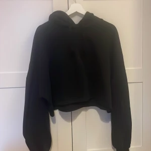 Svart croppad hoodie från Gina Tricot - Svart croppad hoodie från Gina Tricot med huva och långa ärmar. Oversized passform som ger en chill vibe, perfekt till jeans eller mjukisbyxor. Mjuk insida och enkel design utan tryck eller detaljer.