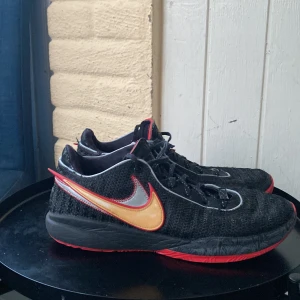 Lebron 20 basketskor/ Svarta Nike sneakers med röd sula - Säljer ett par Lebron 20  , röda detaljer och en stor Swoosh i silver och gult på sidan. Skorna har snörning och en platt, röd sula som ger en sportig vibe. Perfekta för dig som gillar att sticka ut med färg och stil. 