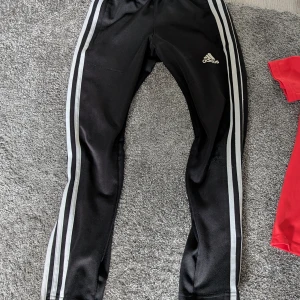 Svarta Adidas träningsbyxor - Svarta träningsbyxor från Adidas med klassiska vita ränder längs sidorna och Adidas-logga på låret. Byxorna har en slim passform och är gjorda i ett glansigt syntetmaterial som passar perfekt för sport och träning.