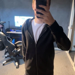 Svart hoodie från Jack & Jones - Svart hoodie från Jack & Jones med dragkedja och huva. Klassisk modell med ribbade muddar och fickor framtill. Perfekt för chill dagar eller när du vill ha en enkel och stilren look. Gjord i mjuk bomullsmix för extra komfort.