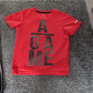 Röd t-shirt med tryck från H&M - Cool röd t-shirt från H&M med stort svart texttryck 'A GAME' framtill. Klassisk rund halsringning och korta ärmar. Perfekt för dig som gillar sportig stil och vill sticka ut med en färgstark look.
