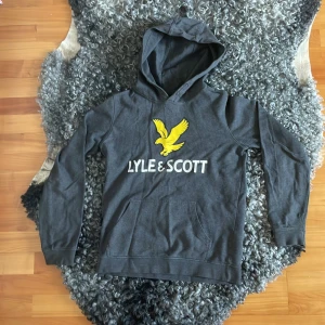 Grå hoodie Lyle & Scott Junior 14/15 år - Mörkgrå hoodie från Lyle & Scott Junior med stor gul logga och text framtill. huva och långa ärmar. Skön och mjuk i bomullsmix.
