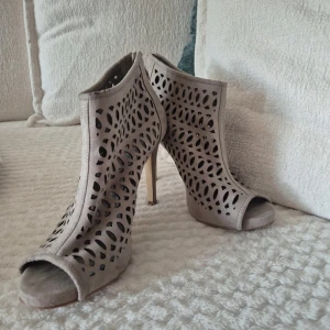 Beige stövletter med öppen tå  - Snygga beige stövletter i mocka-look med coolt utskuret mönster. Skorna har hög klack, öppen tå och dragkedja bak. Perfekta för dig som vill sticka ut med en edgy och trendig stil.