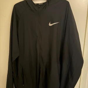Svart vindjacka från Nike med huva - Svart vindjacka från Nike med klassisk logga på bröstet. Jackan har huva, hel dragkedja och två fickor med dragkedja. Perfekt för löpning eller chill dagar. Lätt och smidig design som funkar året runt.