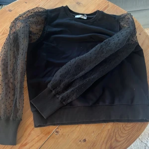 Svart tröja med transparenta ärmar från NAKD - Snygg svart tröja från NAKD med unika transparenta ärmar i prickigt mönster.  Tröjan har en rund halsringning och ribbade muddar vid ärmslut och nederkant.