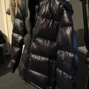 Svart dunjacka från Ralph Lauren i storlek S. - Snygg svart dunjacka från Ralph Lauren med en klassisk pufferdesign. Jackan har en dragkedja framtill och en broderad logotyp på bröstet. Perfekt för kalla dagar. Jackan är i storlek S.