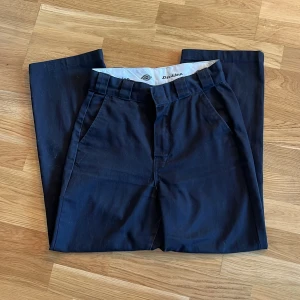 Svarta byxor från Dickies - Snygga svarta byxor från Dickies med en klassisk design. Byxorna har en rak passform och är tillverkade i ett slitstarkt material. Perfekta för en stilren look. De har fickor fram och bak samt en knappstängning.