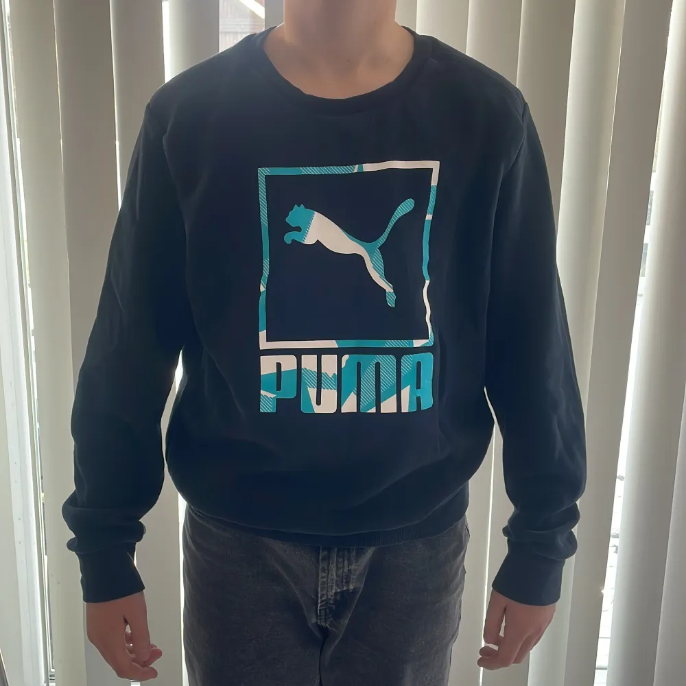 Säljer en mörkblå långärmad tröja från Puma med ett stort logotryck i turkos och vitt på framsidan. Tröjan har en rund halsringning och ribbade muddar vid ärmslut och nederkant. Perfekt för en sportig look ✅. Neuletakit & Villapaidat.