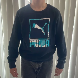 Mörkblå långärmad tröja från Puma - Säljer en mörkblå långärmad tröja från Puma med ett stort logotryck i turkos och vitt på framsidan. Tröjan har en rund halsringning och ribbade muddar vid ärmslut och nederkant. Perfekt för en sportig look ✅