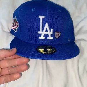 Blå keps från New Era - Snygg blå keps från New Era med LA-logga framtill och broderade detaljer på sidan. Texten 'Love & Hustle = Game' är broderad på sidan i vitt och rött. Perfekt för att ge din outfit en sportig touch.