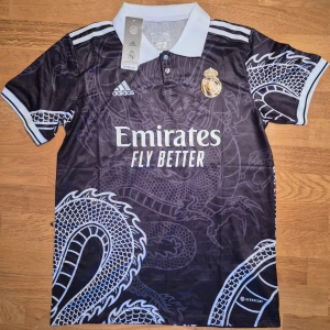 Mbappe tröja Real Madrid - Snygg svart fotbollströja från Adidas med ett coolt drakmönster. Tröjan har korta ärmar och en vit krage. Den har även tryck med 'Emirates Fly Better' och klubbmärke i guld på framsidan. Priset går att diskuteras!