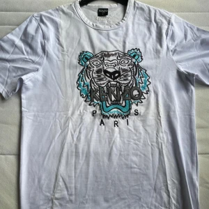 Vit t-shirt med tigerbroderi från Kenzo - Säljer en vit t-shirt från Kenzo med ett coolt broderat tigerhuvud i svart och turkos på framsidan. T-shirten har korta ärmar och en normal passform. Perfekt för att ge din outfit en unik touch!