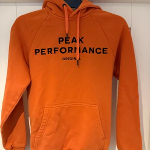 Orange hoodie från Peak Performance - Säljer en snygg orange hoodie från Peak Performance med svart text på framsidan. Tröjan har en klassisk känguruficka och justerbar huva med dragsko. Perfekt för en avslappnad stil.