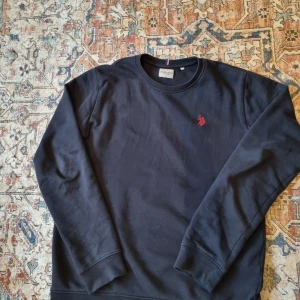 Marinblå tröja från U.S. Polo Assn. - Säljer en stilren svart tröja från U.S. Polo Assn. med ett diskret rött broderi på bröstet. Tröjan har långa ärmar och rund halsringning, perfekt för en avslappnad look.