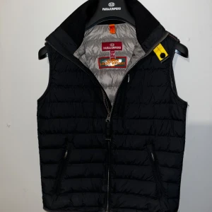 Svart Parajumper Väst - Säljer en svart dunväst från Parajumpers. Västen är superlätt. Den har två fickor med dragkedja och en gul detalj vid kragen. Funkar för höst,vår och sommar🤩 den ända defekten är det lilla hålet som ni kan se på bild 5, det är inget man märker och annars så är västen i mycket bra skick, tags och en galja som man får när man köper finns kvar och den är köpt på kidsbrandstore i malmö. Storlek Y-M alltså barn medium!😁