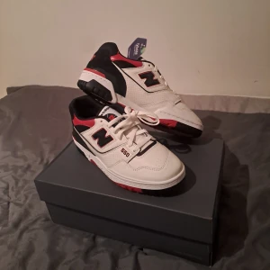 New Balance 550 sneakers - Snygga New Balance 550 sneakers i vitt, svart och rött. Skorna har en klassisk design med snörning och en robust sula i rött och svart. Perfekta för en stilren look.