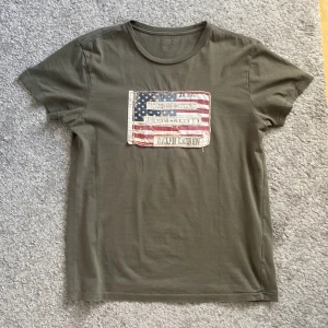 Olivgrön t-shirt från Ralph Lauren - Säljer en olivgrön t-shirt från Ralph Lauren med ett coolt amerikanskt flaggmotiv på bröstet. T-shirten har korta ärmar och en rund halsringning. Perfekt för en avslappnad stil!