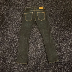 Mörkblåa jeans från Nudie Jeans - Snygga svarta jeans från Nudie Jeans med klassisk femficksdesign och kontrasterande sömmar. Perfekta för en stilren look. Midjan är midwaist och passformen är slim, vilket ger en modern siluett. Hör av er vid minsta fundering🙌