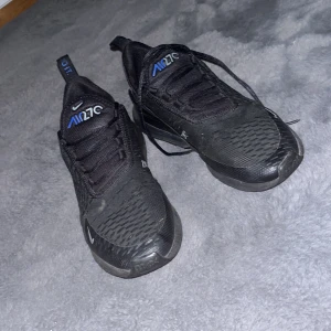 Svarta Nike Air Max 270 - Säljer ett par svarta Nike Air Max 270 med blå detaljer. Skorna har en modern design med synlig Air-enhet i hälen och snörning framtill. Perfekta för både stil och komfort.
