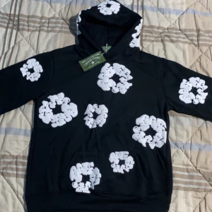 Svart hoodie från Denim Tears - Säljer en svart hoodie från Denim Tears med unikt vitt blommönster över hela plagget. Hoodien har en klassisk passform med en stor ficka framtill och en bekväm huva. Perfekt för en stilren och avslappnad look.