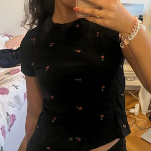 Svart blommig topp - Säljer en svart topp med små röda blommor. Toppen har korta ärmar och en rund halsringning. Perfekt för en avslappnad stil med en touch av blommig charm.