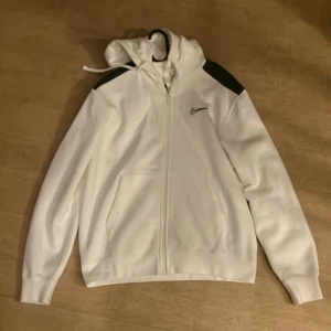 Ziphoodie från Nike  - Säljer en stilren vit hoodie från Nike med dragkedja och gröna axelpartier. Den har en klassisk Nike-logga på bröstet och praktiska fickor framtill. Perfekt för en avslappnad stil.