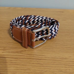 Flätat bälte med läderdetaljer - Snyggt flätat bälte i blått, vitt och brunt med läderdetaljer och silverfärgat spänne. Perfekt för att ge en stilren touch till din outfit.