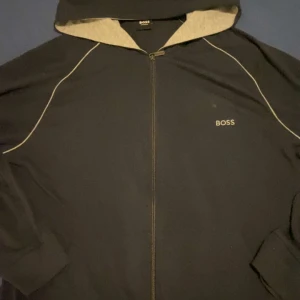 Svart hoodie från Boss - Snygg svart hoodie från Boss med dragkedja och broderad logga på bröstet. Perfekt för en avslappnad stil. Hoodien har en bekväm passform och är idealisk för vardagsbruk.