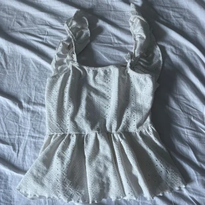 Vit blus med volanger - Säljer en söt vit blus med volanger och spetsdetaljer. Toppen har en öppen rygg med knytband och är ärmlös. Perfekt för sommaren🤍