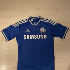 Blå Chelsea fotbollströja från Adidas - Säljer en blå Chelsea fotbollströja från Adidas med korta ärmar och vit text. Tröjan har klubbens emblem på bröstet och Samsung som sponsor. Perfekt för fans av Chelsea FC! 💙2013/14 Kit tror jag. Är lite grön på ryggen och har ett rivmärke på magen.